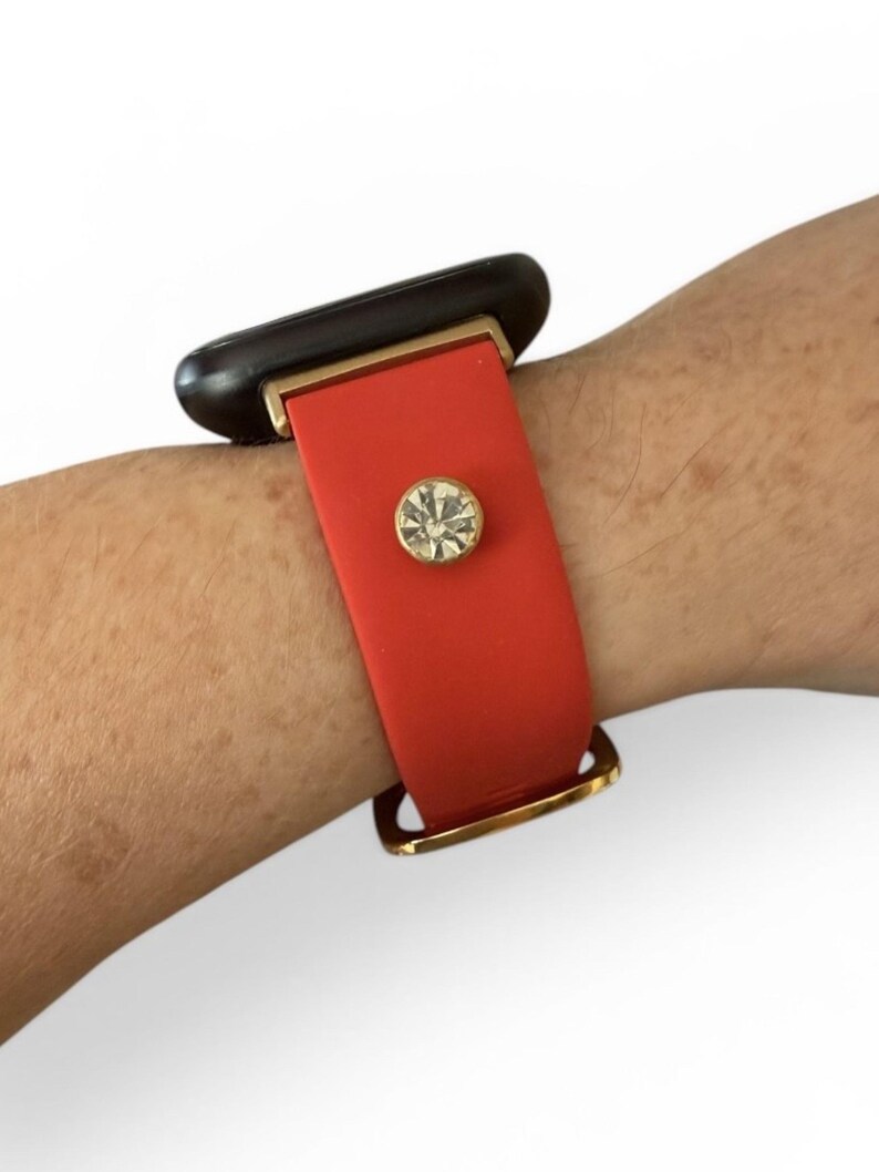 Peut inclure: Un bracelet en silicone rouge avec un accent rectangulaire noir et un fermoir dor&eacute;. Le bracelet est orn&eacute; d'un bijou rond en strass transparent. Le bracelet est port&eacute; sur un poignet.