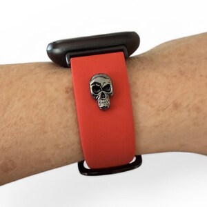 Op de afbeelding: Rode horlogeband met een zilveren schedelbedel. Het horloge is bevestigd aan een zwarte wijzerplaat. De band is gemaakt van een flexibel materiaal.