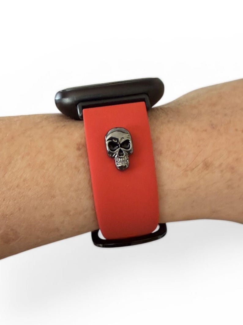 Op de afbeelding: Rode horlogeband met een zilveren schedelbedel. Het horloge is bevestigd aan een zwarte wijzerplaat. De band is gemaakt van een flexibel materiaal.