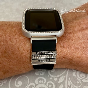 K&ouml;nnte beinhalten: Eine silberne Smartwatch mit einem schwarzen Silikonarmband und einer mit Kristallen besetzten L&uuml;nette. Das Armband hat drei silberne Charms mit den eingravierten W&ouml;rtern "Blessed", "Faith" und "Hope".