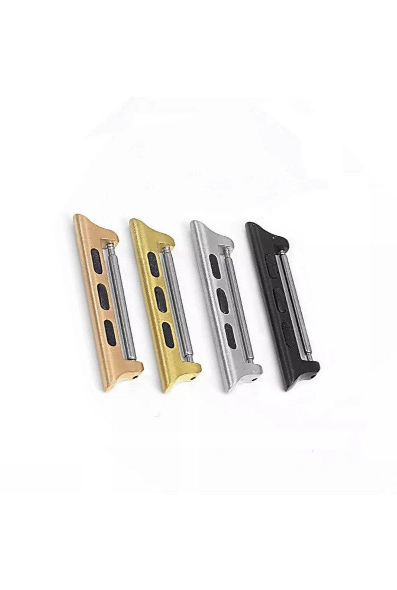 May include: Four watch band adapters in gold, yellow gold, silver, and black. Each adapter has a rectangular shape with rounded edges and a metal bar for attaching to a watch band. The adapters are angled slightly.
