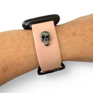 Op de afbeelding: Een lichtroze smartwatchband met een zwarte sluiting en een gunmetal schedelbedel. De band is bevestigd aan een zwart smartwatch-apparaat. De schedelbedel is gecentreerd op de band.