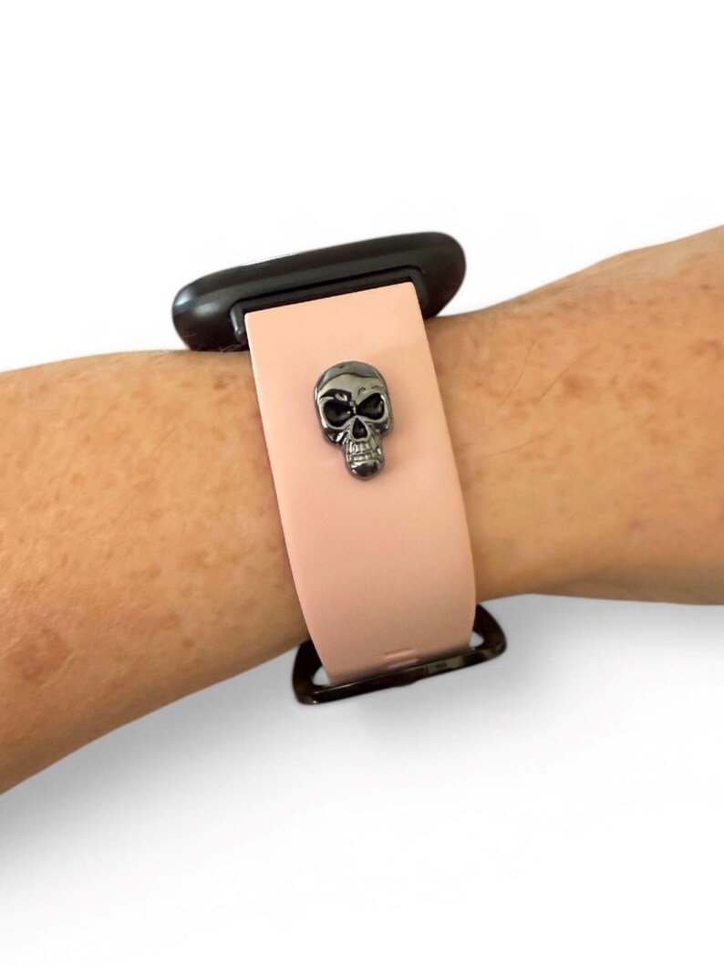 Op de afbeelding: Een lichtroze smartwatchband met een zwarte sluiting en een gunmetal schedelbedel. De band is bevestigd aan een zwart smartwatch-apparaat. De schedelbedel is gecentreerd op de band.