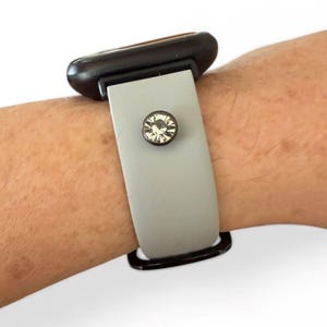Peut inclure: Un bracelet de montre gris avec un fermoir noir et un cristal clair, rond et d&eacute;coratif. Le bracelet est attach&eacute; &agrave; un cadran de montre noir.