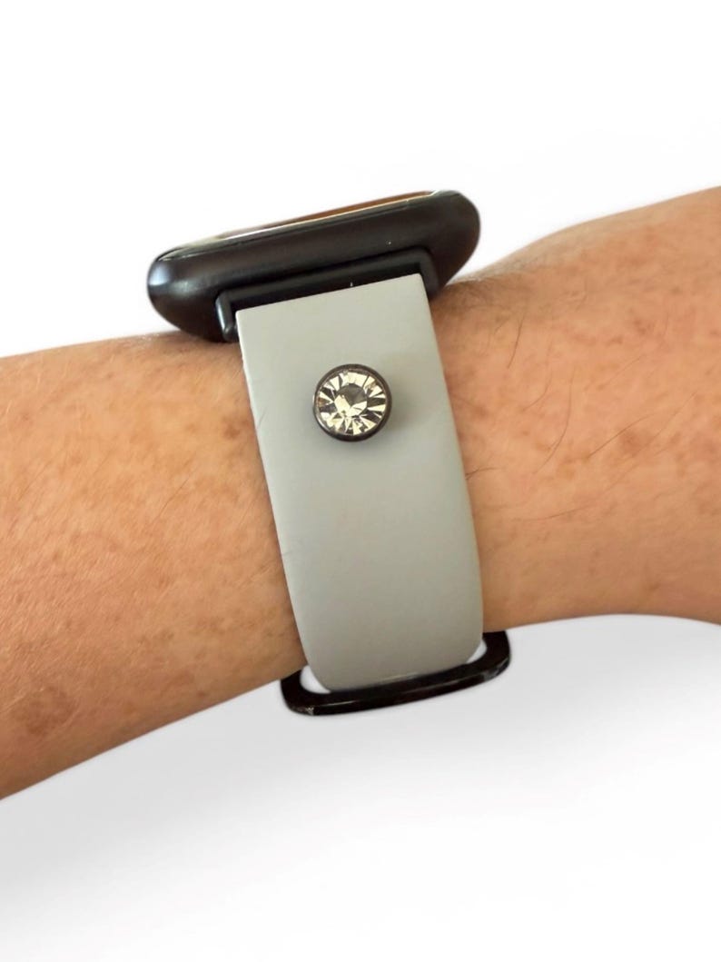 Peut inclure: Un bracelet de montre gris avec un fermoir noir et un cristal clair, rond et d&eacute;coratif. Le bracelet est attach&eacute; &agrave; un cadran de montre noir.