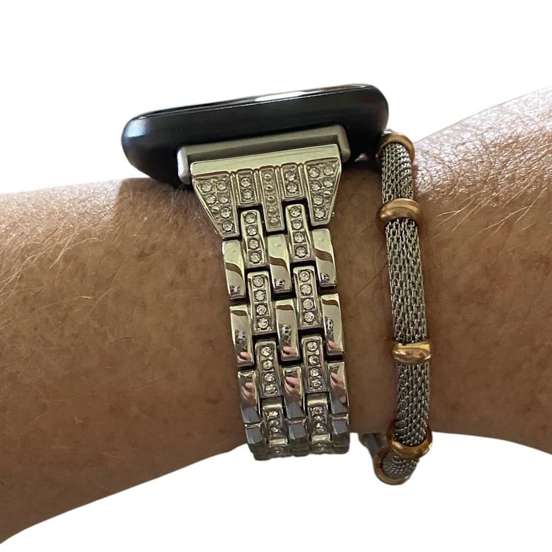 May include: Silver-toned watch band with a rectangular link design, embellished with clear rhinestones. The watch face is black. A silver and gold mesh bracelet is next to the watch band.