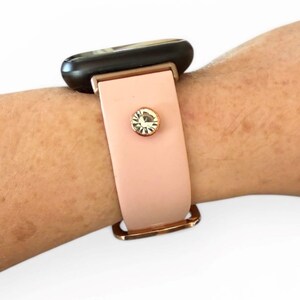 Peut inclure: Un bracelet de montre rose clair avec une boucle dor&eacute;e et un cristal d&eacute;coratif. Le cadran de la montre est noir. Le bracelet est attach&eacute; &agrave; un poignet.