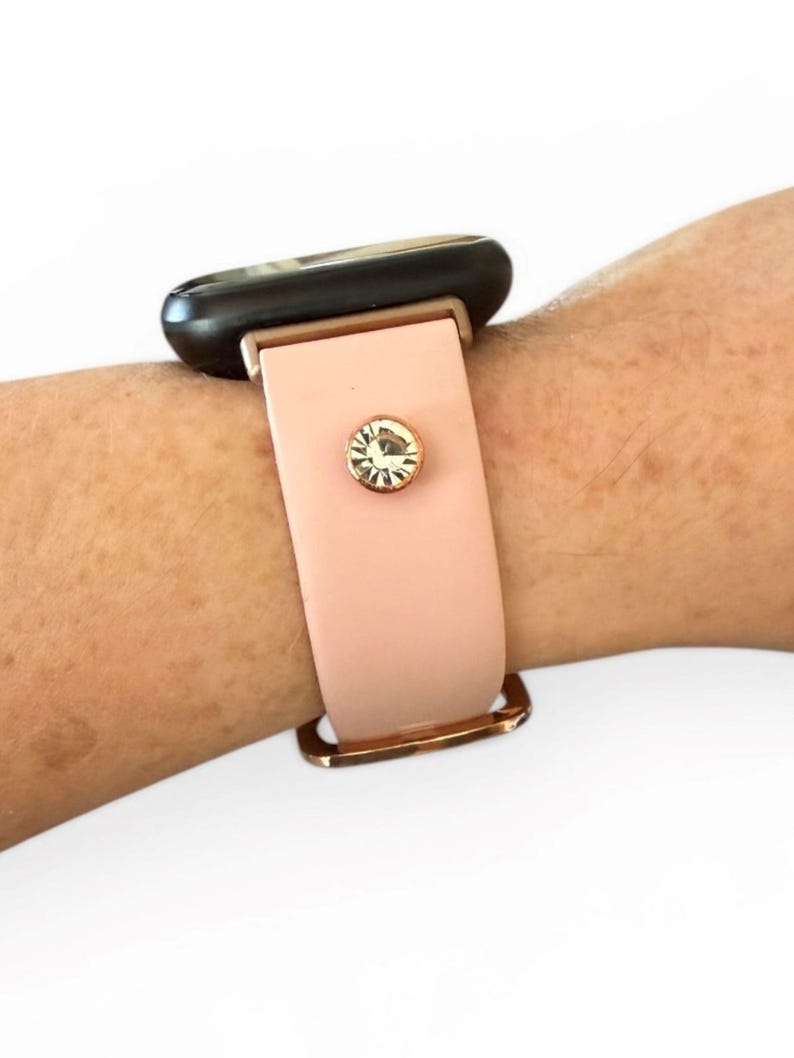 Peut inclure: Un bracelet de montre rose clair avec une boucle dor&eacute;e et un cristal d&eacute;coratif. Le cadran de la montre est noir. Le bracelet est attach&eacute; &agrave; un poignet.