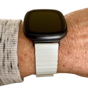 Peut inclure: Un bracelet de montre en silicone blanc avec une montre intelligente noire. Le cadran de la montre est noir et rectangulaire.
