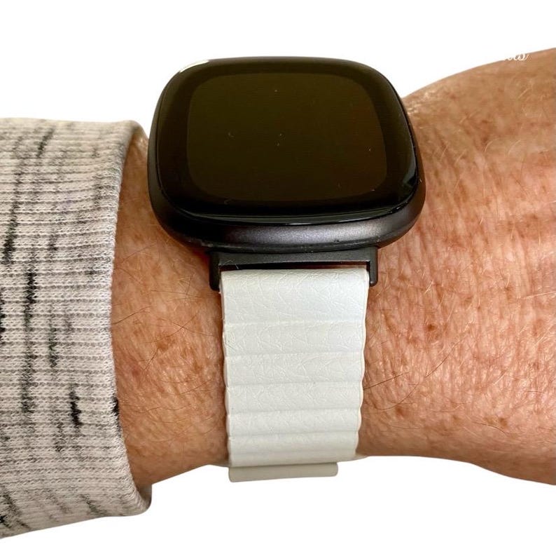 Peut inclure: Un bracelet de montre en silicone blanc avec une montre intelligente noire. Le cadran de la montre est noir et rectangulaire.