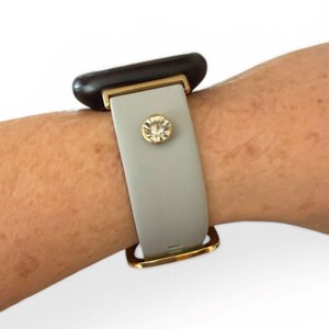 Peut inclure: Un bracelet de montre intelligente gris clair avec des accents dor&eacute;s et un embellissement en cristal &eacute;tincelant. Le cadran de la montre est noir. Le bracelet est fix&eacute; avec un fermoir dor&eacute;.