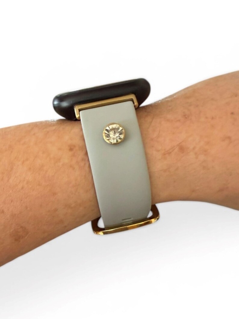 Peut inclure: Un bracelet de montre intelligente gris clair avec des accents dor&eacute;s et un embellissement en cristal &eacute;tincelant. Le cadran de la montre est noir. Le bracelet est fix&eacute; avec un fermoir dor&eacute;.