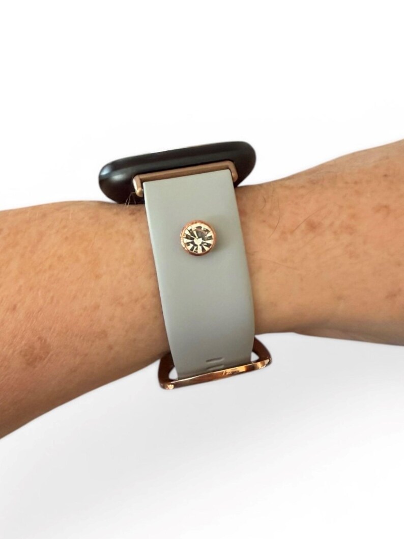 Peut inclure: Un bracelet de montre gris clair avec une boucle en or rose et un accent d&eacute;coratif en cristal. Le cadran de la montre est noir. Le bracelet est fix&eacute; au poignet.