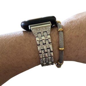 May include: Silver-toned Apple Watch with a rhinestone-studded band. A mesh bracelet with gold accents is worn alongside the watch. The watch has a black face and is displayed on a wrist.