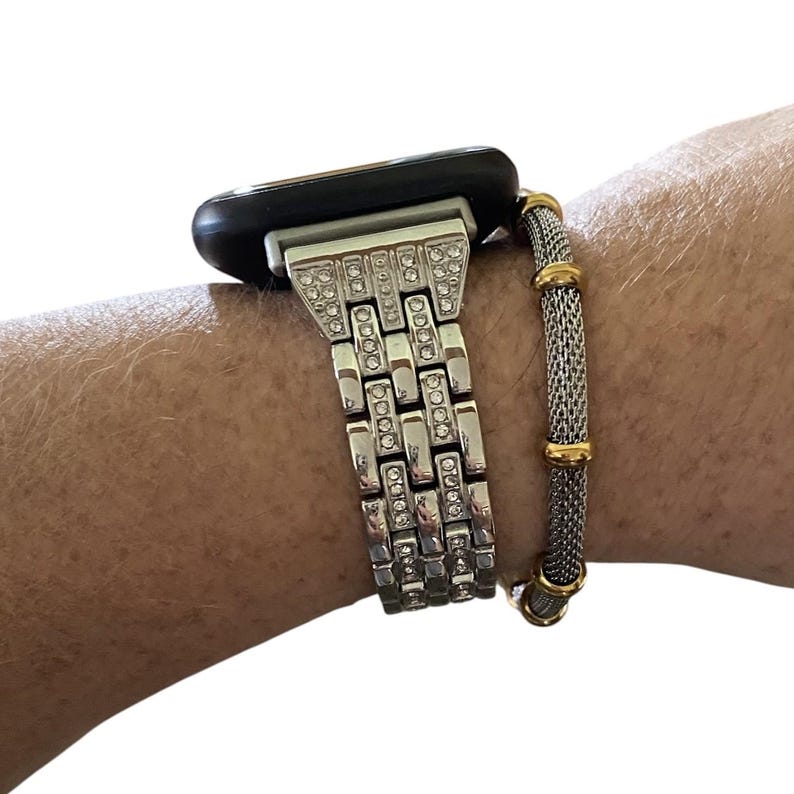 May include: Silver-toned Apple Watch with a rhinestone-studded band. A mesh bracelet with gold accents is worn alongside the watch. The watch has a black face and is displayed on a wrist.