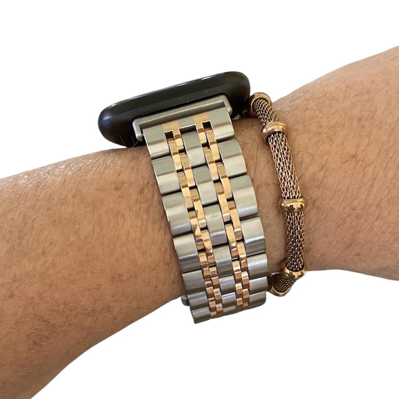 Peut inclure: Un bracelet de montre argent&eacute; et dor&eacute; est attach&eacute; &agrave; une montre intelligente noire. Un bracelet en maille dor&eacute;e s&eacute;par&eacute; est port&eacute; &agrave; c&ocirc;t&eacute; du bracelet de montre. Le bracelet de montre est compos&eacute; de maillons argent&eacute;s avec des accents dor&eacute;s.