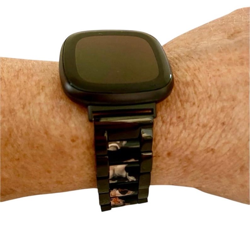 Puede incluir: Un reloj inteligente negro con una banda de metal negro con un patr&oacute;n floral. La esfera del reloj es negra y rectangular.