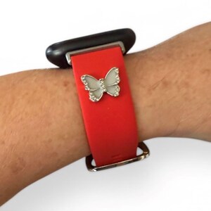 May include: Red smartwatch band with a silver butterfly charm. The butterfly is embellished with small, clear crystals. The band is secured with a silver buckle and is attached to a black smartwatch face.