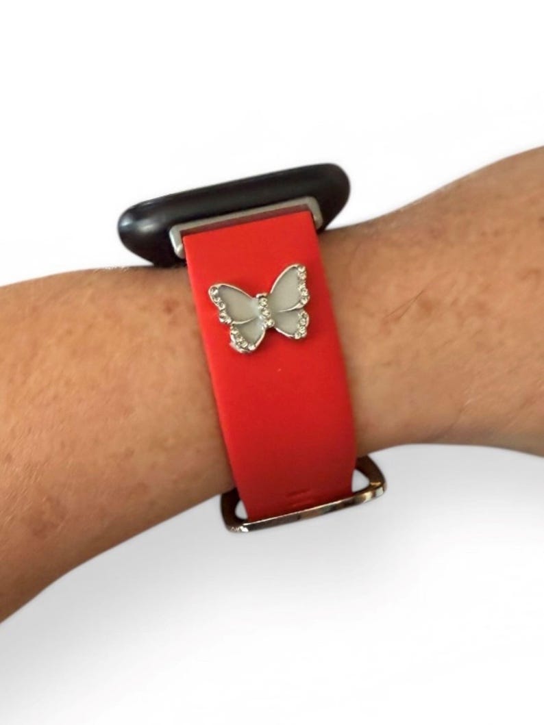 May include: Red smartwatch band with a silver butterfly charm. The butterfly is embellished with small, clear crystals. The band is secured with a silver buckle and is attached to a black smartwatch face.