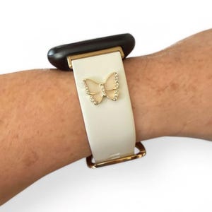 May include: White watch band with a gold-colored butterfly charm. The band has gold-colored metal accents and is attached to a black watch face. The butterfly charm is embellished with small, clear crystals.