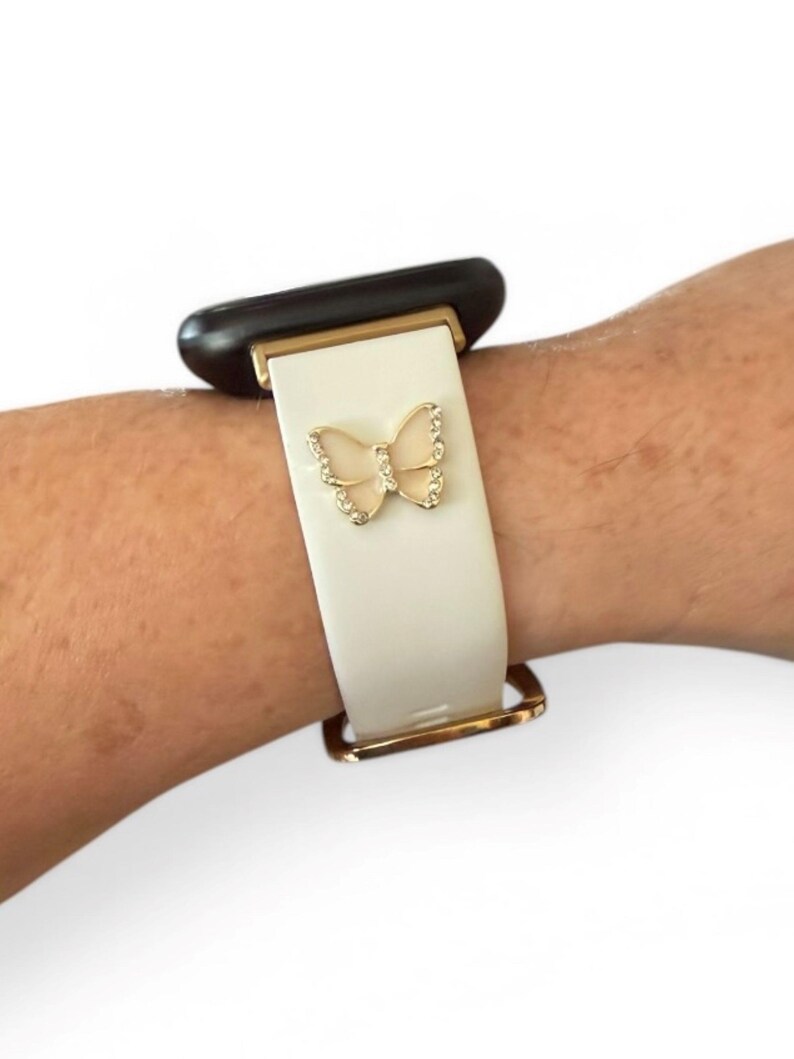 May include: White watch band with a gold-colored butterfly charm. The band has gold-colored metal accents and is attached to a black watch face. The butterfly charm is embellished with small, clear crystals.