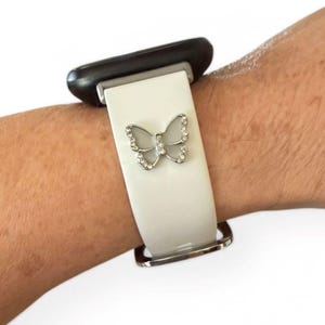 May include: A white watch band with a silver butterfly charm. The butterfly is adorned with small, clear crystals. The watch band is attached to a black watch face and a silver clasp.