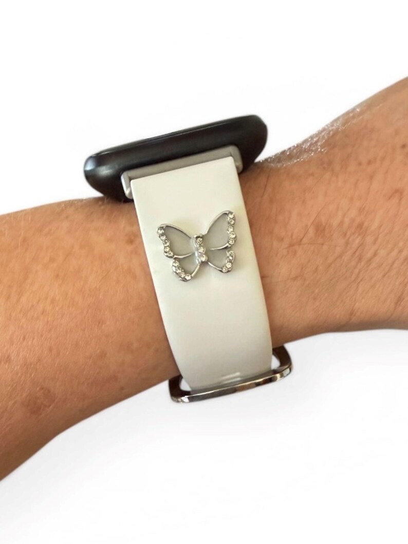 May include: A white watch band with a silver butterfly charm. The butterfly is adorned with small, clear crystals. The watch band is attached to a black watch face and a silver clasp.