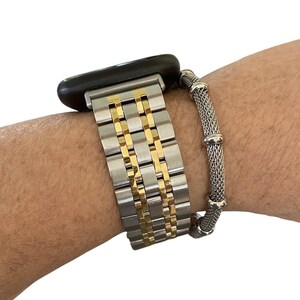 Peut inclure: Un bracelet de montre argent&eacute; et dor&eacute; est attach&eacute; &agrave; une montre intelligente noire. Un bracelet en maille argent&eacute;e s&eacute;par&eacute; est &eacute;galement visible. Le bracelet de montre a un design &agrave; maillons rectangulaires avec des accents dor&eacute;s.
