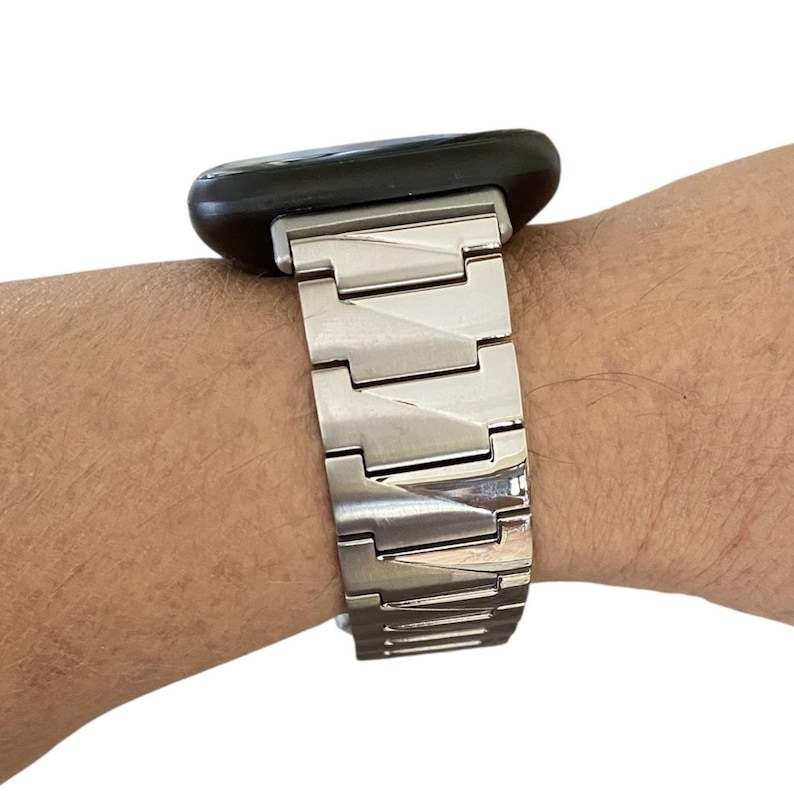 K&ouml;nnte beinhalten: Ein silbernes Uhrenarmband mit einem schwarzen Zifferblatt wird an einem Handgelenk gezeigt. Das Uhrenarmband besteht aus Metallgliedern. Das Zifferblatt ist rechteckig mit abgerundeten Ecken. Das Uhrenarmband ist am Zifferblatt befestigt.