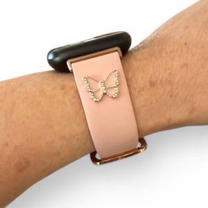 May include: A light pink watch band with a gold-colored buckle and a decorative butterfly. The butterfly is embellished with small, clear crystals. The watch face is black.