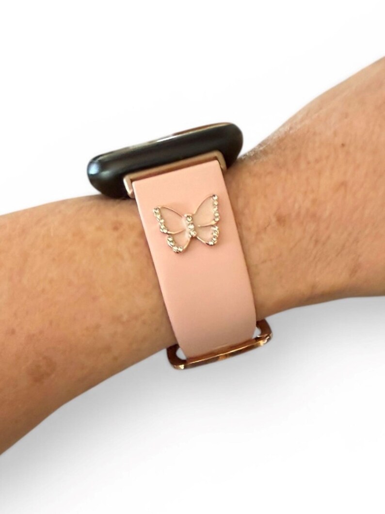 May include: A light pink watch band with a gold-colored buckle and a decorative butterfly. The butterfly is embellished with small, clear crystals. The watch face is black.