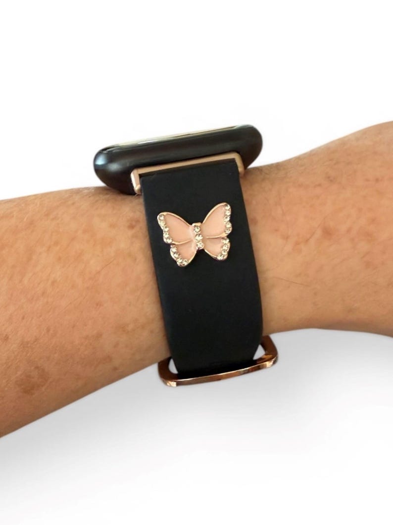 May include: Black watch band with a rose gold buckle and a pink butterfly charm. The butterfly is embellished with small, clear crystals. The watch face is black.