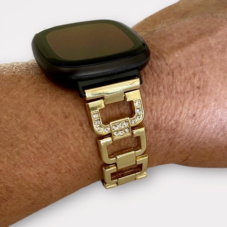 May include: Gold watch band with clear rhinestones. The band is made of metal links and has a square shape. The band is designed to fit a smartwatch.