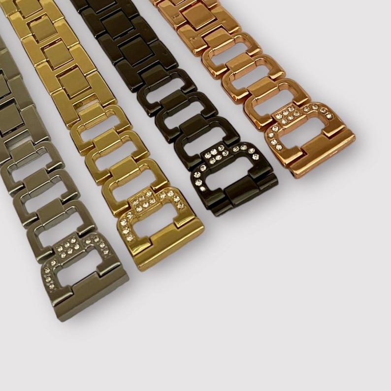 May include: Four watch bands with a linked chain design. The bands are in gold, rose gold, black, and silver. Each band has a row of clear rhinestones.