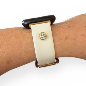 Peut inclure: Un bracelet de montre blanc avec une boucle et des accents dor&eacute;s. Le bracelet est orn&eacute; d'un grand cristal rond et transparent. Le cadran de la montre est noir.