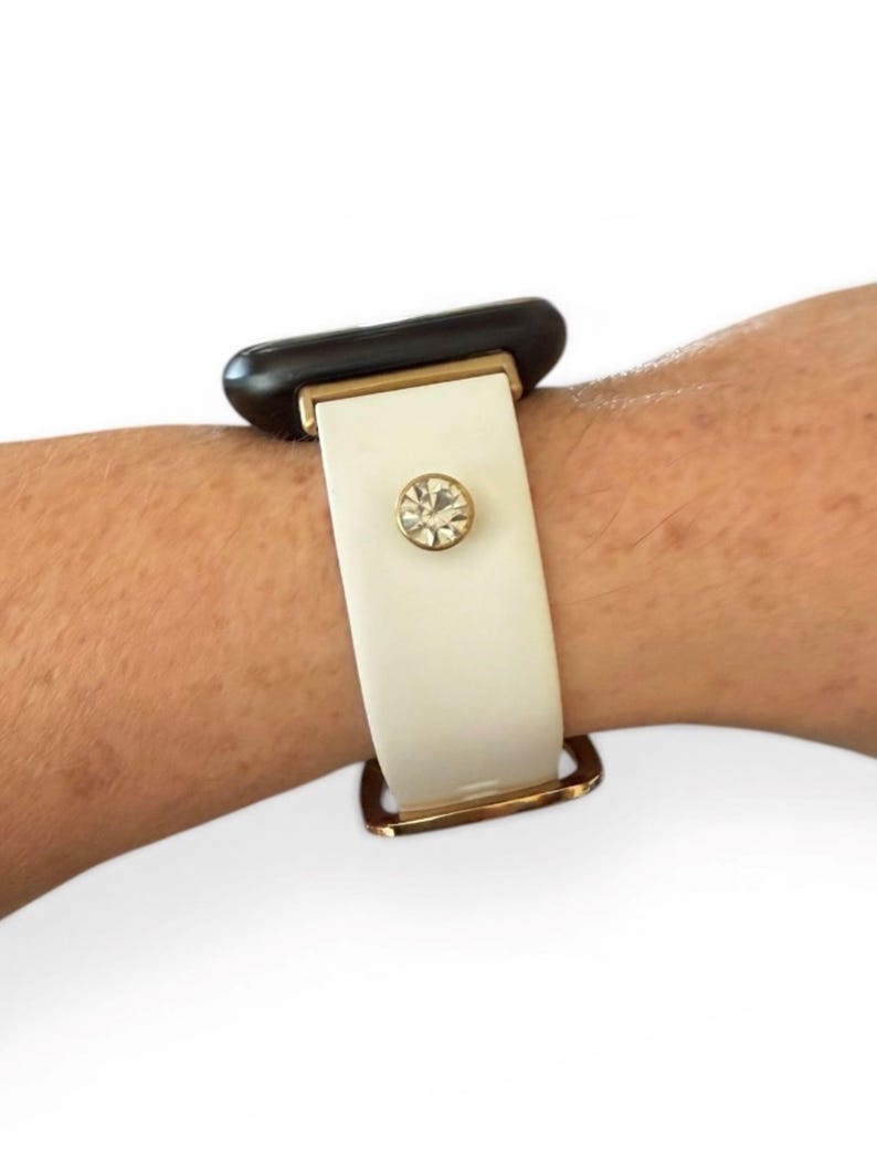 Peut inclure: Un bracelet de montre blanc avec une boucle et des accents dor&eacute;s. Le bracelet est orn&eacute; d'un grand cristal rond et transparent. Le cadran de la montre est noir.