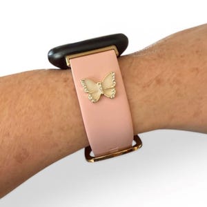 May include: A pink smartwatch band with a gold-colored buckle and a decorative butterfly. The butterfly is embellished with small, clear crystals. The watch face is black.
