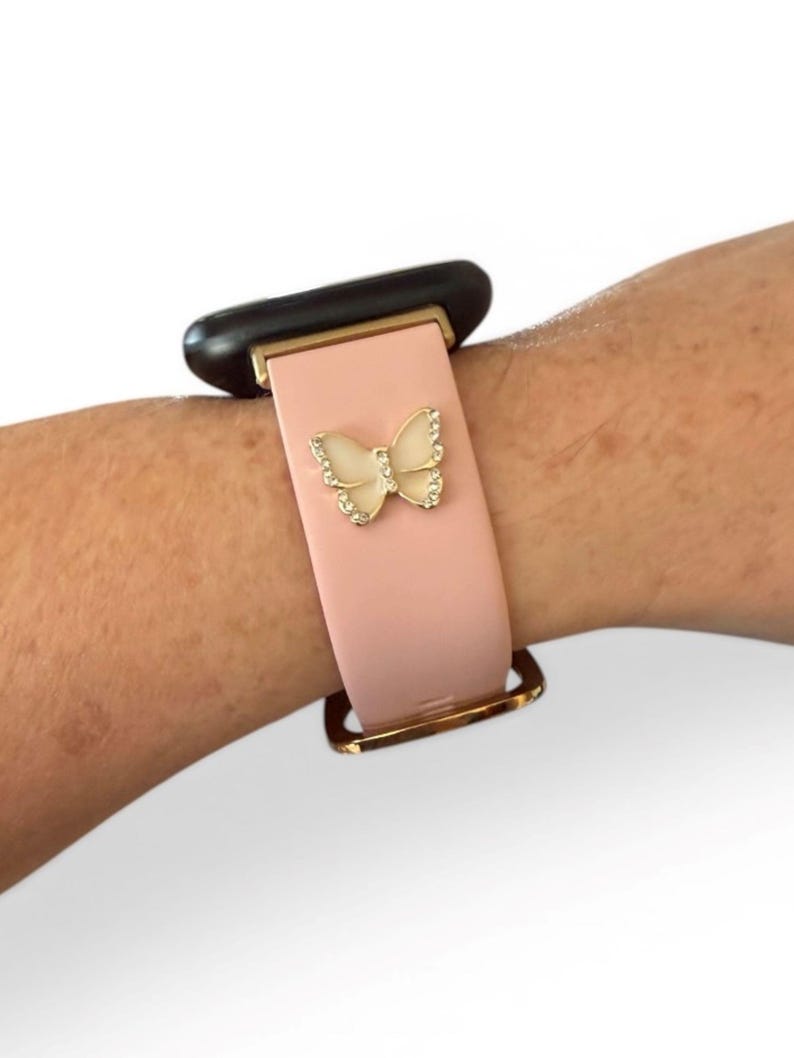 May include: A pink smartwatch band with a gold-colored buckle and a decorative butterfly. The butterfly is embellished with small, clear crystals. The watch face is black.