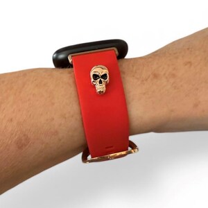 Op de afbeelding: Rode smartwatchband met een gouden schedelbedel. De band is bevestigd aan een zwart smartwatch-apparaat. De band is vastgezet met een goudkleurige gesp. De schedelbedel is gecentreerd op de band.
