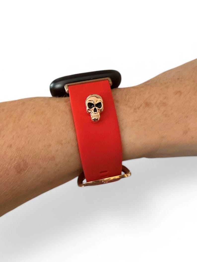 Op de afbeelding: Rode smartwatchband met een gouden schedelbedel. De band is bevestigd aan een zwart smartwatch-apparaat. De band is vastgezet met een goudkleurige gesp. De schedelbedel is gecentreerd op de band.