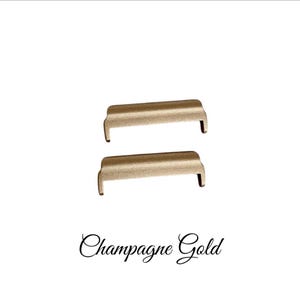May include: Two gold-toned metal watch band strap adapters. The adapters are rectangular with rounded edges. The text "Champagne Gold" is written below the adapters.