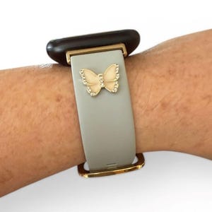 May include: A gray smartwatch band with a gold-colored butterfly charm. The butterfly is embellished with small, clear crystals. The band has gold-colored metal accents and is attached to a black watch face.