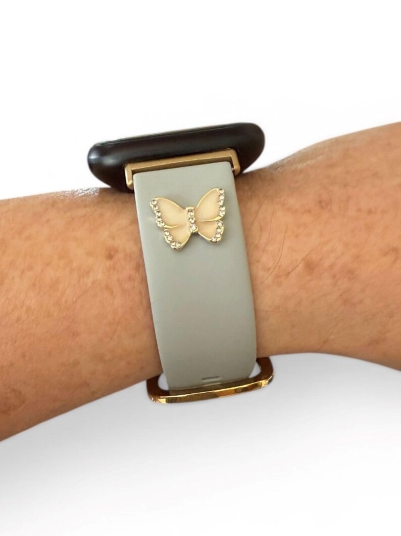 May include: A gray smartwatch band with a gold-colored butterfly charm. The butterfly is embellished with small, clear crystals. The band has gold-colored metal accents and is attached to a black watch face.