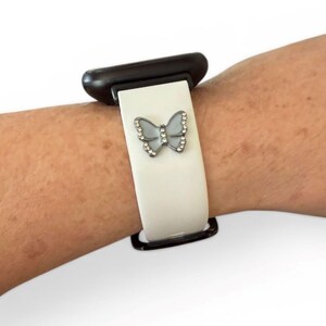May include: A white watch band with a decorative butterfly charm. The butterfly is silver-toned and adorned with small, clear crystals. The watch band is attached to a black watch face.