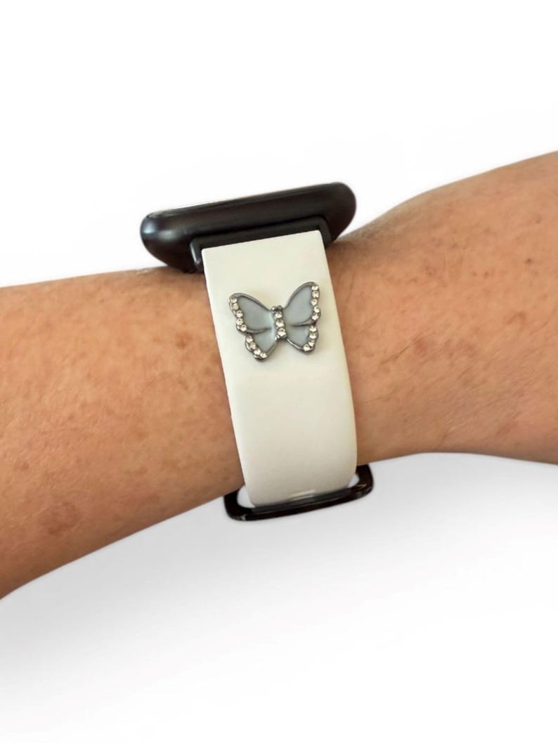 May include: A white watch band with a decorative butterfly charm. The butterfly is silver-toned and adorned with small, clear crystals. The watch band is attached to a black watch face.