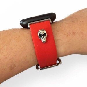 Op de afbeelding: Rode horlogeband met een zilveren schedelbedel. De horloge is bevestigd aan een zwarte wijzerplaat. De band is vastgezet met een zilveren gesp.