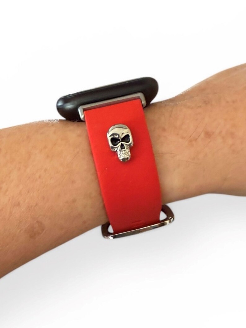 Op de afbeelding: Rode horlogeband met een zilveren schedelbedel. De horloge is bevestigd aan een zwarte wijzerplaat. De band is vastgezet met een zilveren gesp.