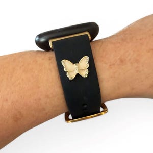 May include: Black watch band with a gold-colored buckle and a decorative butterfly. The butterfly is cream-colored with gold trim and small, clear rhinestones. The watch face is dark brown.