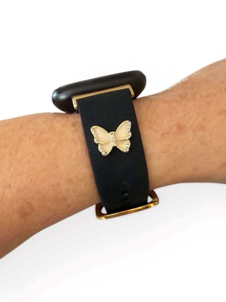 May include: Black watch band with a gold-colored buckle and a decorative butterfly. The butterfly is cream-colored with gold trim and small, clear rhinestones. The watch face is dark brown.