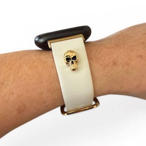 Op de afbeelding: Witte horlogeband met een gouden schedelbedel. De band heeft goudkleurige hardware en is bevestigd aan een zwarte wijzerplaat.